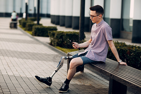 有脚假肢的残疾青年男子坐在户外手持手机和在户外安放移动电话图片