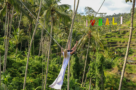 一个年轻的女孩在高高的峡谷上荡秋千 巴厘岛热带丛林雨林的悬崖上荡秋千的年轻女游客 秋千悬挂在两棵椰子树之间 我爱巴厘岛天空吸引力图片