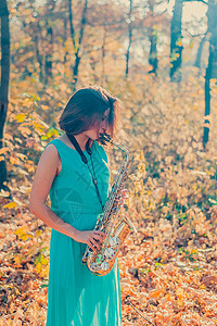 穿着长蓝色裙子的深褐色女孩 在秋森林里演黄色萨克斯风剧背景图片