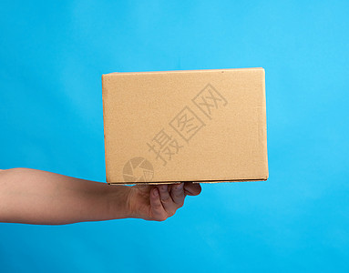 手手里拿着一盒蓝色背景的棕色纸纸箱导游店铺邮政盒子货运纸盒正方形材料货物纸板图片