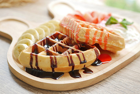 配上草莓糖浆和巧克力酱的美味华夫饼 边盘菜是新鲜草莓切碎 香蕉 奶油和冰淇淋 放在可爱木板上图片
