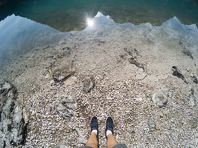穿着鞋子的人腿站在湖水线和地面附近 湖中有山的倒影 在晴天 白云岩阿尔卑斯山 意大利 欧洲 在运动相机上拍摄图片