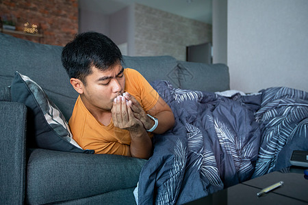 亚洲男子在家里沙发上生病和咳嗽 他用温度计检查冠状病毒流行期间的温度 10月1日过敏疾病鼻子喷嚏感染男性药品客厅症状病人图片