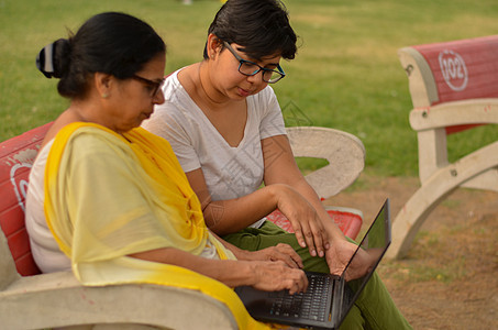 在印度新德里的一个公园里 年轻的印度女孩在坐在红色长椅上的笔记本电脑上帮助一位印度老妇人的侧视图 概念数字素养教育老年公民人士孙图片