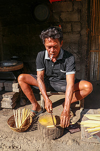 在印度尼西亚巴厘岛 老家伙在做竹草的车间里砍竹子 前面看一个制作竹草的工人生态力量日志金属工具柳条建筑曲线工作男性图片