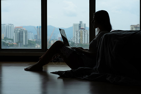 Siluet 女孩在笔记本电脑上工作 喝着咖啡 坐在靠近全景窗户的床边的地板上 时尚现代的内饰 舒适的工作场所 在互联网上购物互图片
