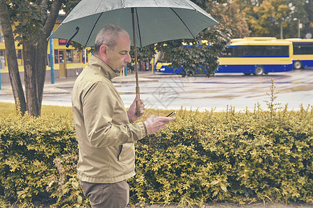 一个男人拿着雨伞站在公交车站等车 男人在等公共汽车 在雨天用手机发短信 当地小镇的雨天图片