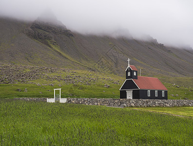 绿色草地上的黑木红屋顶教堂 雾中的陡峭山 Raudisandur 冰原西峡湾图片