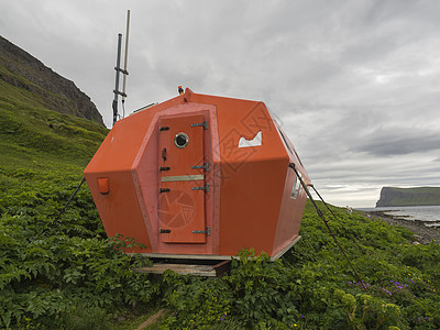 的红色紧急避难所小屋站在海边草草地上 在冰岛西码头角城悬崖旁观图片