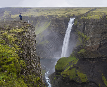 冰岛南部的 Fossa 河谷和美丽的 Haifoss 瀑布 身穿蓝色夹克的男子站在悬崖岩石上看风景图片