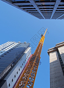 在周围有大型塔楼的新混凝土建筑上工作的黄色高大建筑起重机的垂直视图图片