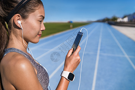 跑步音乐激励女人戴着耳机听电话应用程序 赛跑者看着体育场跑道上的智能手机 耳机和手机准备好运行 健康的生活方式图片