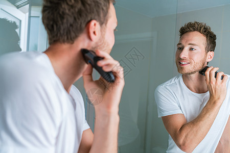 在浴室用电动剃须刀剃须 男人照镜子 男性美容常规生活方式图片