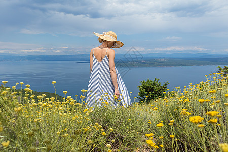 身穿条纹夏季服装和草帽的年轻女子站在野花开花丛露出 放轻松而美观亚得里亚海自然的近视中 克罗地亚太阳阳光冥想海滩天空生活幸福喜悦图片