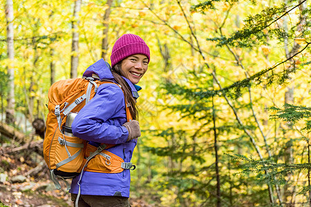 快乐的背包客女孩在秋天的森林里徒步旅行 年轻的亚洲徒步旅行者女性穿着户外装备以应对寒冷的天气 背着背包看着相机 在秋季享受在大自图片