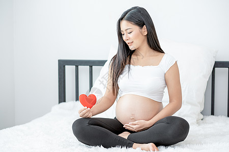 美丽的亚洲孕妇的肖像 坐在床上并举着红心图标保健婴儿微笑家庭母亲卧室母性女士卫生产妇图片