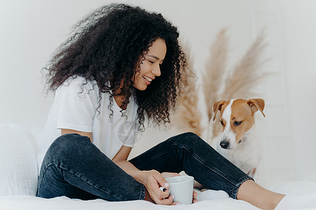 可爱的卷发女狗主人的照片看着动物微笑 拿着一杯茶 坐在白色宽敞卧室的床上 表达对动物的爱 人 宠物和友谊概念图片