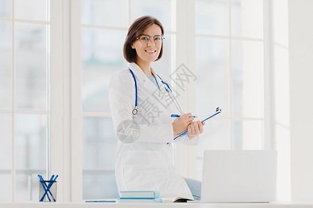 专业女医生的室内照片提供医疗护理协助 在台式机上摆姿势 带笔记本电脑 准备文件协议纸报价 戴光学眼镜和白色医疗礼服图片