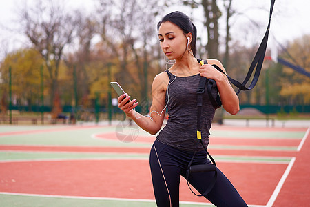 在运动场上使用 trx 进行女运动员训练 混血年轻成年女性使用悬挂系统进行锻炼 健康的生活方式 伸展户外游乐场女士微笑耳机运动公图片