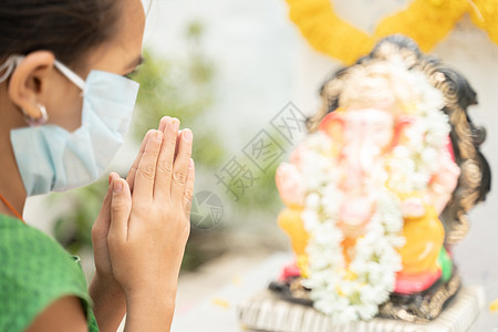 在 Ganesha 或节期间 戴着医用面具的女孩在 Ganesha 勋爵面前闭上眼睛祈祷——covid-19 或冠状病毒大流行期图片