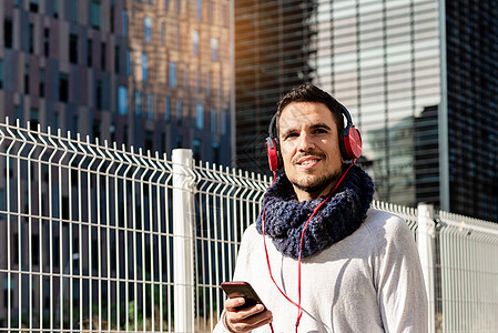 阳光明媚的日子里 留着胡子的年轻男性戴着耳机 手持智能手机 在摩天大楼前行走城市电话围巾音乐微笑街道晴天技术享受企业家图片