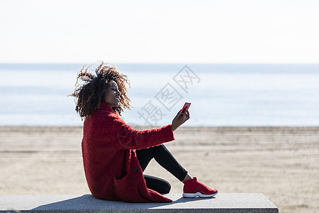 在阳光明媚的一天 使用手机坐在岸边的美美美美女女士女孩微笑发型日光休闲装爆炸成人长椅海滩图片