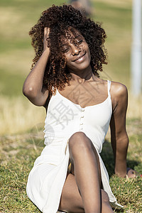 阳光明媚的日子里 身穿白色裙子的美丽非洲裔美国女性坐在公园的草地上成人微笑日光女孩地面女士休闲装潮人发型卷曲图片