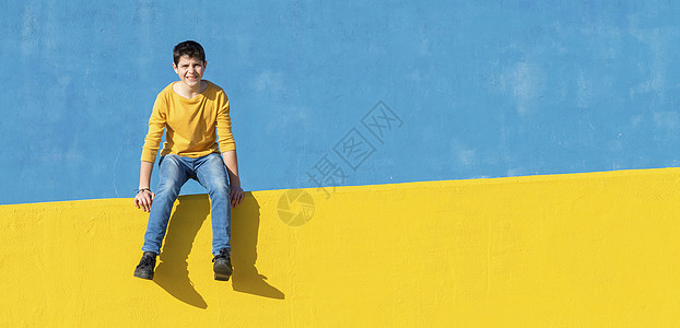 阳光明媚的日子里 一个穿着休闲服的小男孩坐在黄色的篱笆上 靠着蓝色的墙壁太阳日光牛仔裤男生城市衣服男性街道青少年男人图片