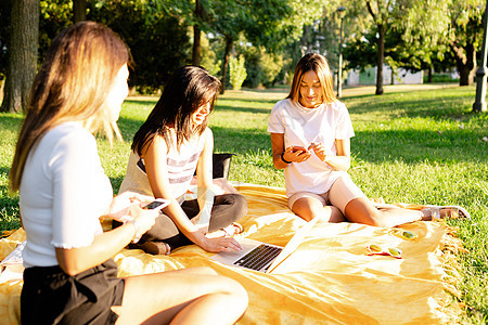 三位美丽的年轻白人女性朋友早上坐在公园的草地上使用智能手机 — 人们的新技术习惯 — 在户外使用互联网进行智能工作图片