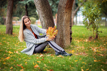 一个穿灰色羊毛衫的漂亮女孩坐在草地上 手里握着秋天黄叶花束图片