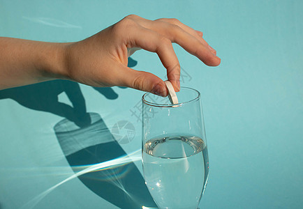 在蓝色背景下 一只手将一片溶解的冻状阿司匹林药片投进一杯水中玻璃图片