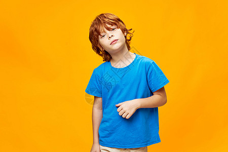 红头发男孩手握着一件蓝色T恤衫 将头部倾斜到黄色孤立背景的一边图片