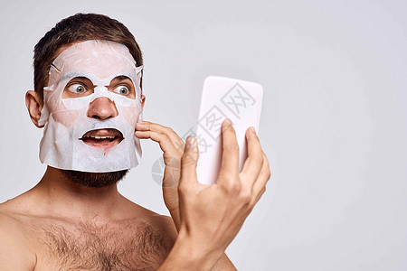 一个脸上戴清洗面罩的男子 在光背景上用镜子照自己的脸部头发浴室卫生剃须治疗护理眼睛微笑男生男人图片