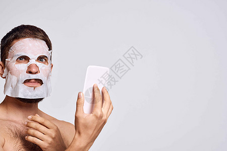 一个脸上戴清洗面罩的男子 在光背景上用镜子照自己的脸部卫生奶油微笑成人男生治疗身体浴室眼睛头发图片