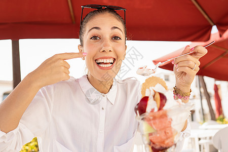 美丽的微笑快乐的年轻白人女性一边用手指做着美味的手势 一边吃着一个带水果的大复合冰淇淋 — 暴食者新的不良饮食习惯图片