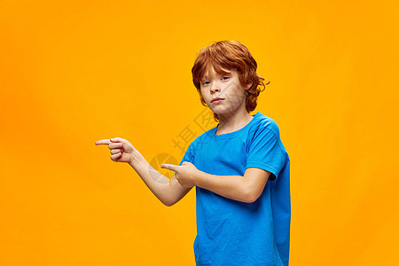 一个穿着蓝色T恤衫的红色头发男孩 向侧面看指数指 黄色孤立背景图片