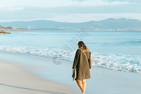 一位穿着大衣的年轻女子 在秋天阳光明媚的白天 在海滩上岸边举着他的鞋子图片