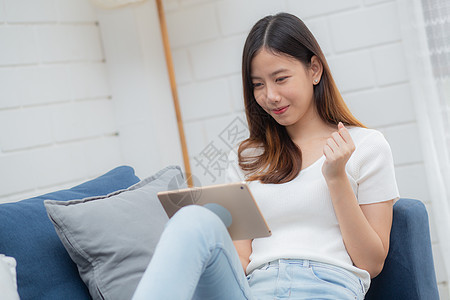 年轻迷人的亚洲女性在家里的沙发上使用浏览平板电脑休息 快乐的女孩坐在沙发上放松阅读数字小工具 在家里兴奋 交流和生活方式概念自由图片