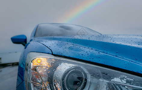 蓝色豪华 SUV 汽车前视图在雨天停在海边附近 彩虹为背景 汽车引擎盖蓝色纹理上的雨滴 汽车在恶劣天气下打开前照灯以确保安全驾驶图片