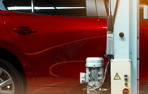 汽车修理厂有选择地关注红色闪亮的汽车和带电机和电机控制箱的升降机杆 红色 SUV 汽车在车库中提升以进行维修和保养服务 汽车侧面图片