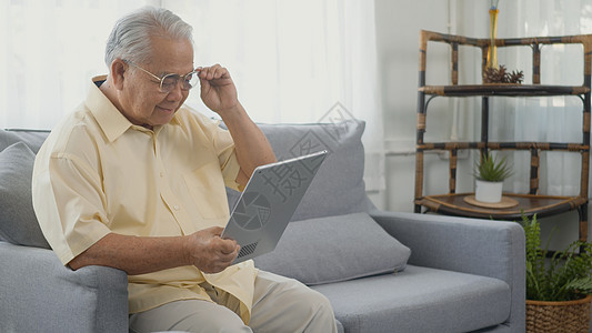 在沙发上坐着穿着眼镜戴眼镜的年长男子在数字平板电脑上寻找消息图片