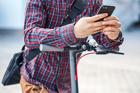 年轻男子身穿衬衣 靠着电动摩托车车把手栏 手里握着移动智能手机 特写工具潮人城市生态衬衫运输细胞电话车辆街道图片