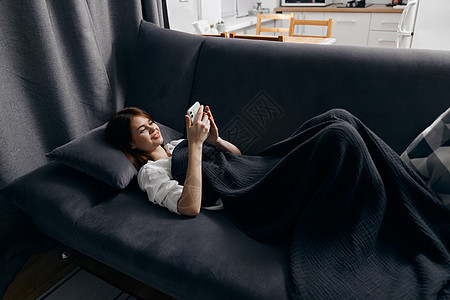 一个带手机的女人 躺在一个灰色的沙发上 在背景中靠近窗户的窗边图片
