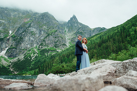 年轻一对夫妇在环绕山峰的湖边散步夫妻绿色幸福成人婚礼会议眼镜后台新娘支撑图片