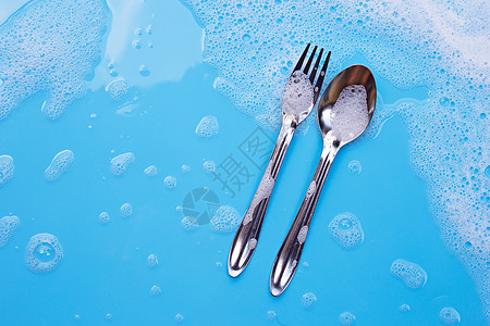 洗碗概念 用肥皂泡沫在湿蓝背景上用汤匙叉子和香皂泡沫图片