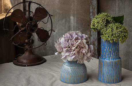 阳台房旧水泥墙白色质感桌布上的两朵蓝色手工陶瓷花瓶和复古棕色金属扇中的花束图片