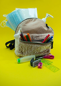 学校用品 如铅笔剪刀橡皮尺水彩面罩酒精消毒剂 放在黄色背景的棕色袋子背包里 在带有复制空间的 covid 流行病期间回到学校概念图片