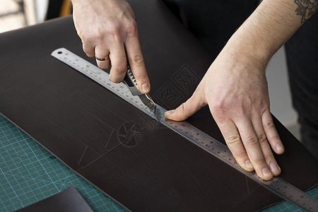 男人的手拿着文具刀和金属尺子 在他的车间里切割皮革钱包的碎片 与棕色天然皮革的工作过程 拿着制作工具的工匠手工产品机器工艺配饰维图片