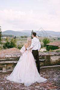 在意大利托斯卡纳的一座古老的酒庄别墅举行婚礼 一对新婚夫妇站在一家老酒厂的屋顶上 拥抱在一起 背对着画框站立金发女郎女士发型羽毛图片