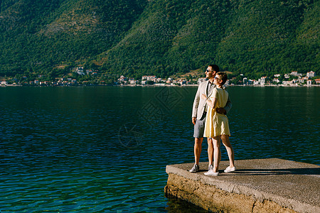 一对相爱的情侣站在Perast附近的Kotor湾的一个码头上拥抱着绿色斜坡的背景图片